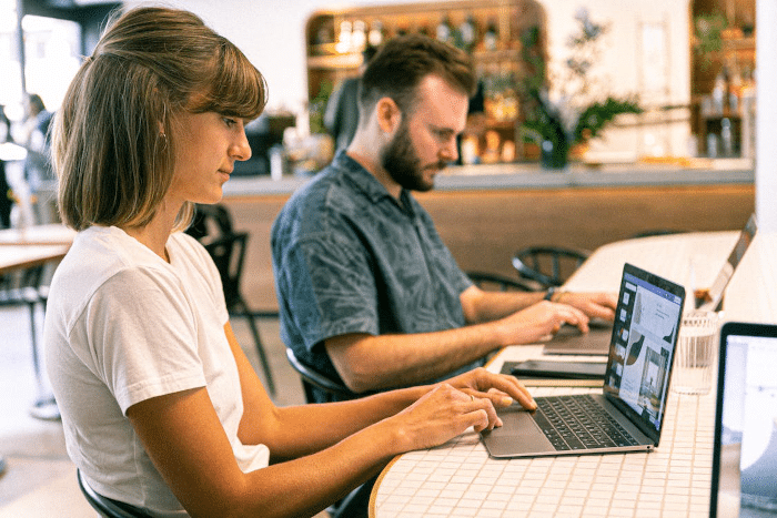 Eine Frau und ein Mann sitzen in einem Café nebeneinander und haben ihre Blicke auf die Bildschirme ihrer Laptops gerichtet.